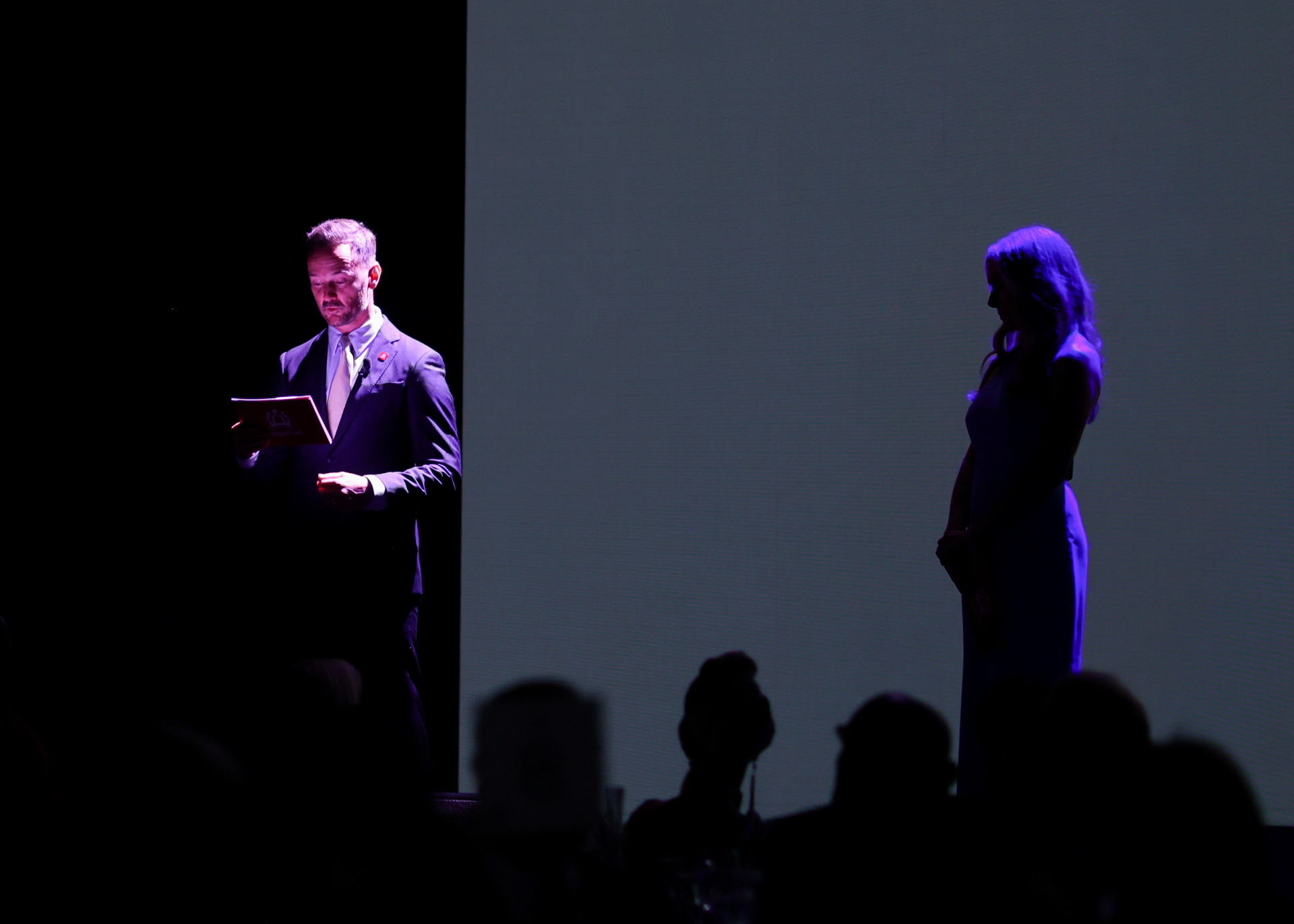 Michael Welch reading from a programme in a spotlight on stage at The King's Trust gala, the audience in silhouette