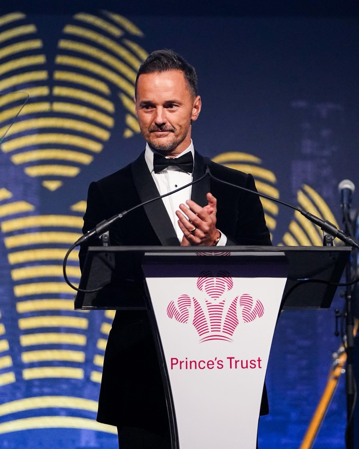 Michael Welch in black tie, applauding at a lectern bearing The Prince's Trust logo