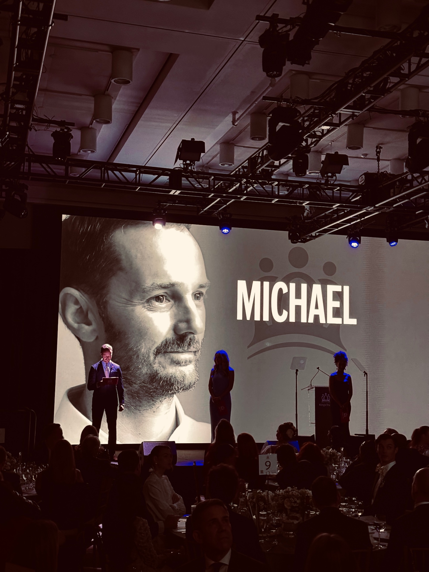 A King's Trust event stage, with a large portrait of Michael Welch and his first name projected on screen behind the speakers